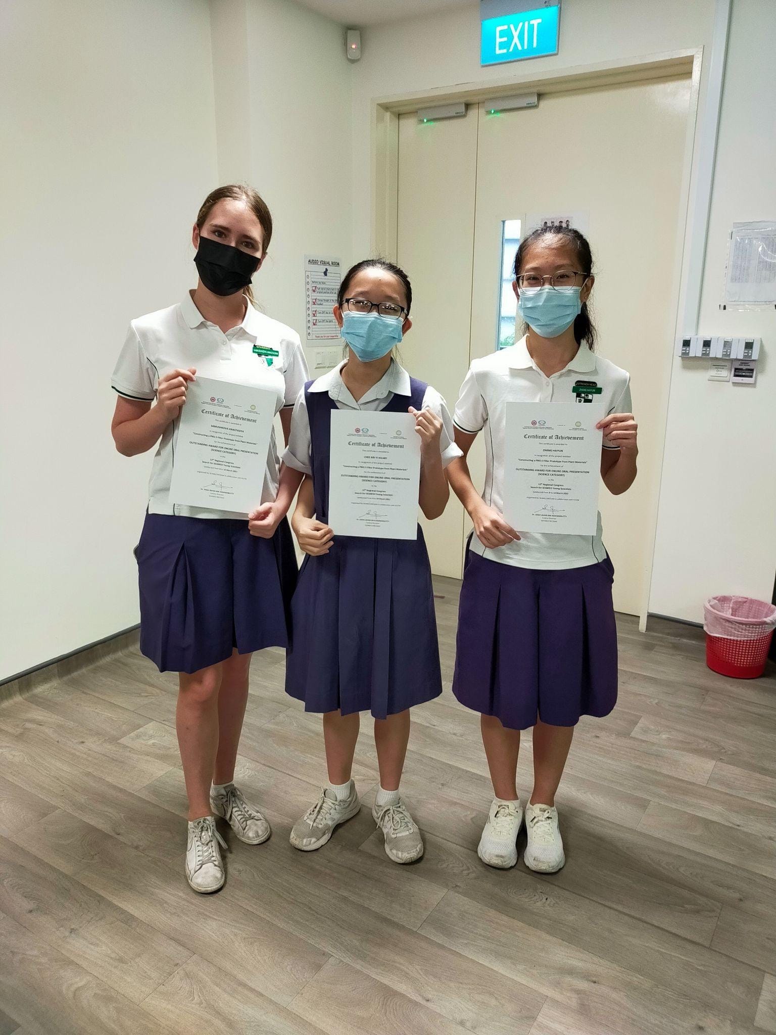 Three masked schoolgirls in white and purple uniforms hold certificates of achievement indoors near an exit door.